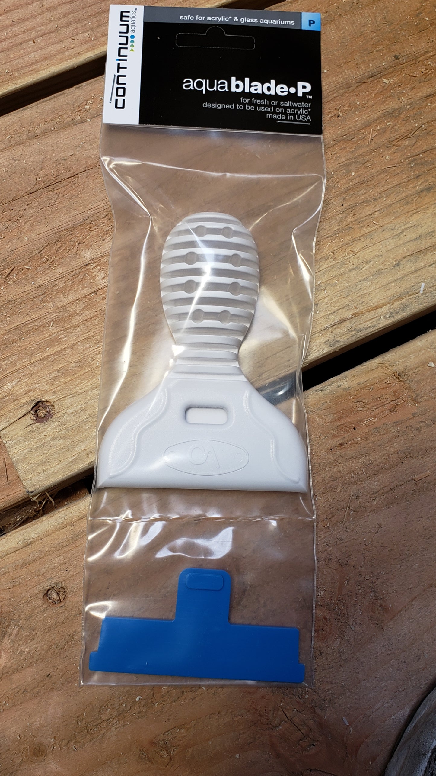 An aquarium scraper with a white and blue acrylic blade, packaged in a clear plastic bag with a label reading 'AquaBlade P', set against a wooden background.