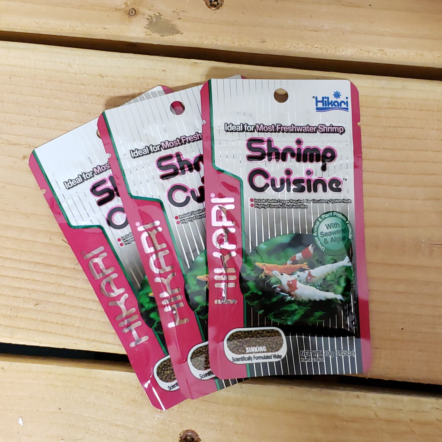 Three packages of Hikari Shrimp Cuisine Pellets placed on a wooden surface.